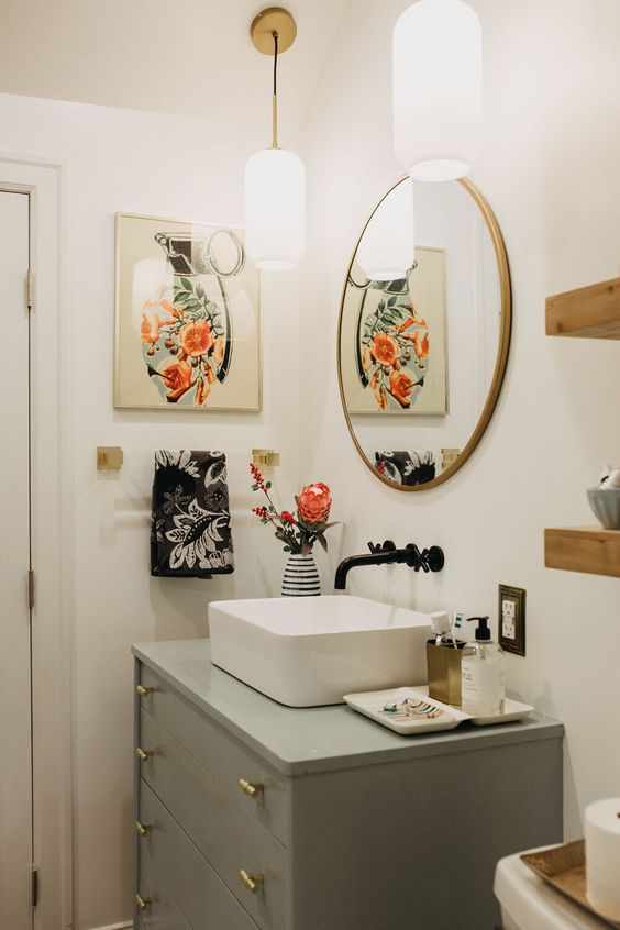an eclectic bathroom with a light grey vanity, a round mirror, a bold artwork, wooden shelves and a sink with a black faucet