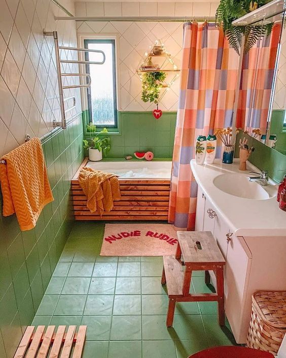an eclectic bathroom with a white and green tiles, a tub clad with wood, a pink vanity and a bold curtain and orange towels