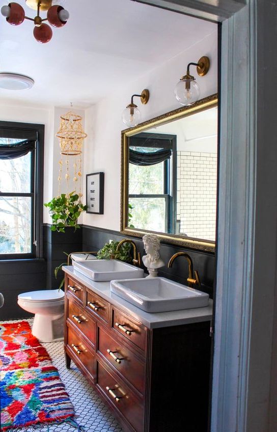 an eclectic bathroom with black paneling, a black window frame, a stained vanity, a large mirror in a gilded frame, a bold rug and sconces