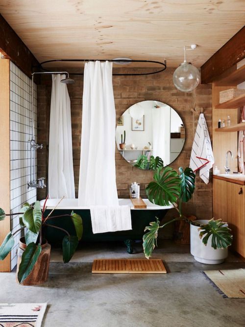 an eclectic bathroom with brick walls, a stone floor, a black free-standing bathtub, potted plants and a round mirror