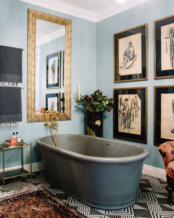an eclectic bathroom with light blue walls, a large gallery wall, a stone tub, a tile floor, a mirror in a gilded frame