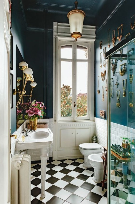 an eclectic bathroom with navy walls and a gallery wall of jewelry, a vintage free-standing sink, glam lamps and gold accessories