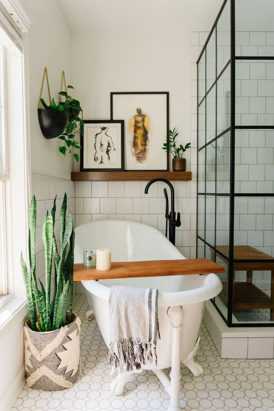 an eclectic bathroom with white square tiles, a free-standing tub,  a shower space, a shelf with art and potted greenery