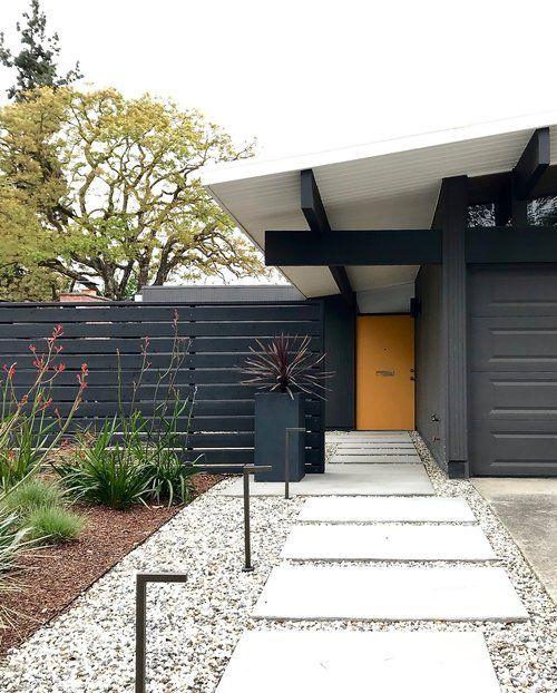 an elegant modern front yard with gravel and large tiles, greenery and blooms gorwing next to the black fence and a tall planter