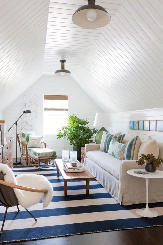 a small attic and beachy living room done with white shiplap on the walls and ceiling, neutral furniture and a striped rug