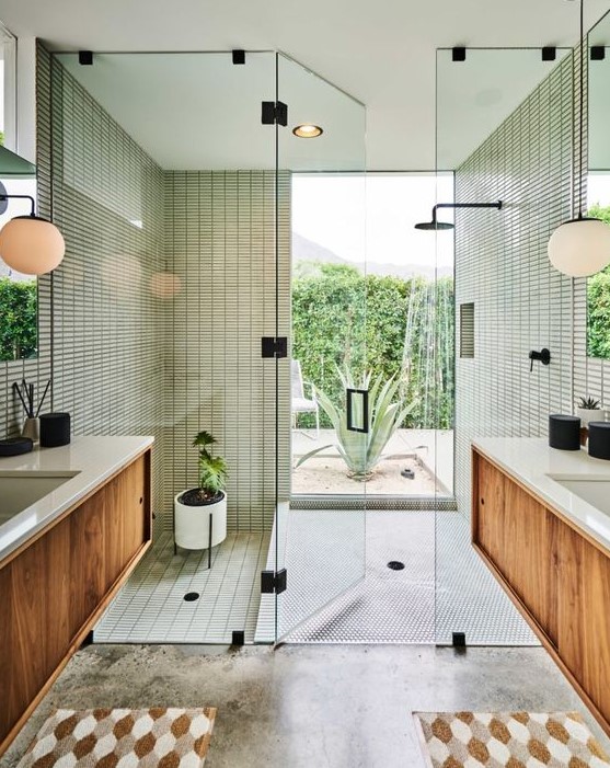 a mid-century modern bathroom with skinny green tiles in the shower and a window, stained vanities, printed rugs and potted plants