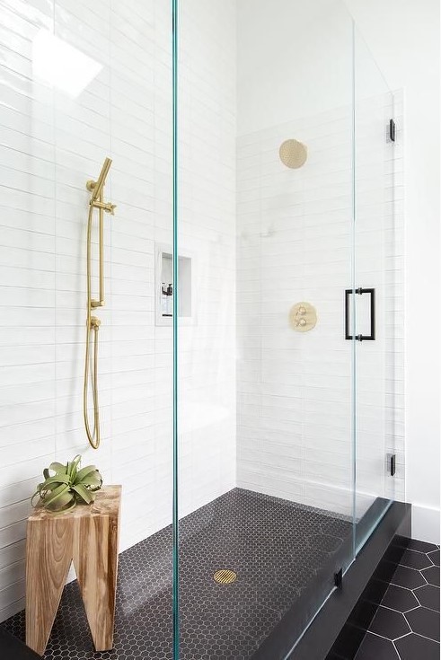 a mid-century modern bathroom with stacked white tiles, black penny and hexagon ones, a wooden stool and brass touches