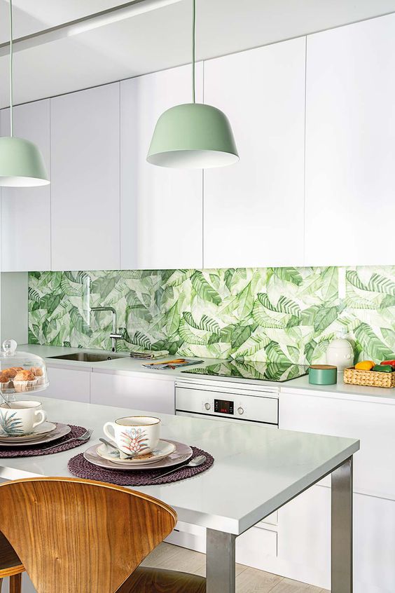 a modern tropical kitchen with sleek white cabinets, a tropical leaf print backsplash, green pendant lamps and wooden chairs