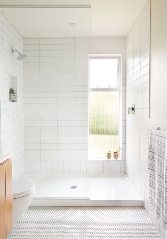 a neutral bathroom with stacked white tiles in the shower, penny tiles on the floor, a light-stained floating vanity and a window