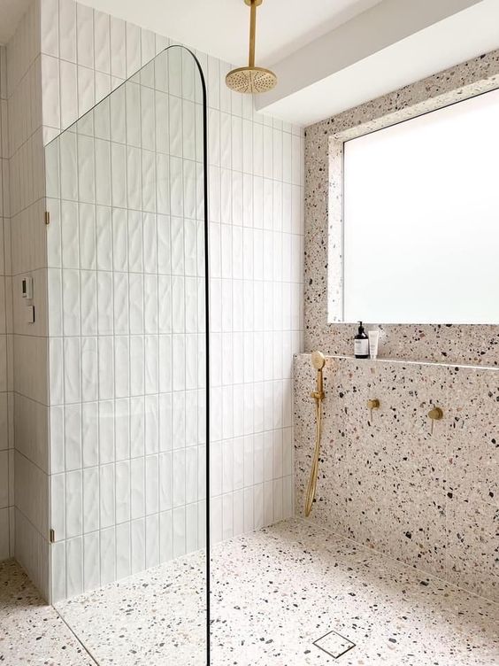 a neutral shower space with neutral skinny tiles and catchy terrazzo, gold fixtures and a window is a lovely space to take a shower