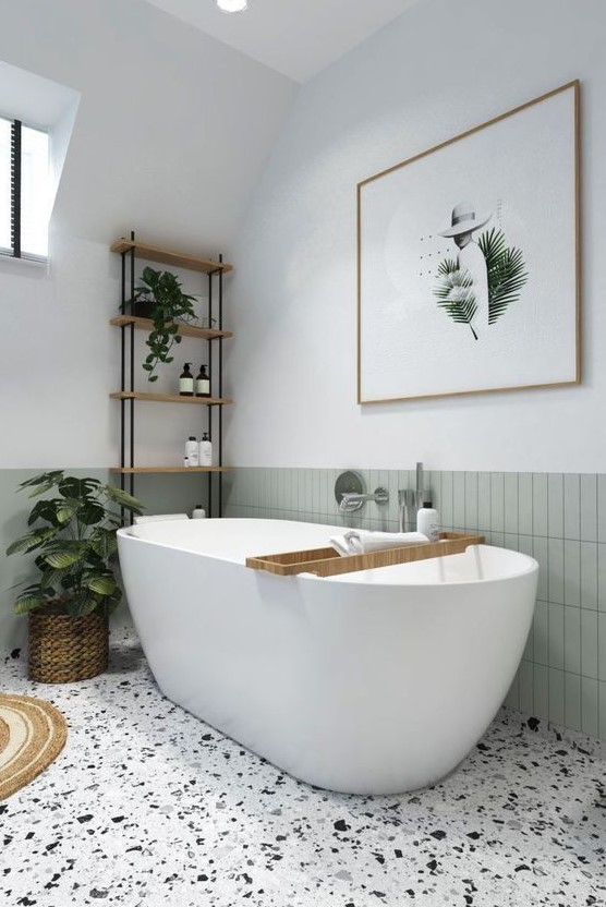 a serene neutral bathroom with a light green tile stacked backsplash, a terrazzo floor, an oval tub, potted plants and a bold artwork