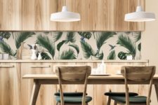 a simple and laconic tropical kitchen with plywood cabinets, a tropical leaf backsplash, a wooden table and upholstered chairs