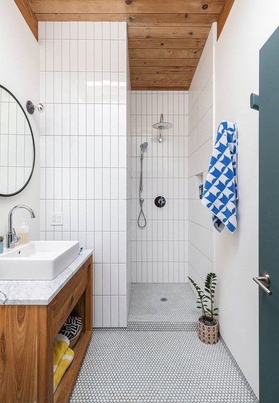a simple mid-century modern bathroom with white penny and skinny tiles, a wooden vanity and ceiling and a round mirror
