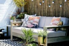a small deck with a large comfortable sofa, lights over it and lots of potted greenery and blooms