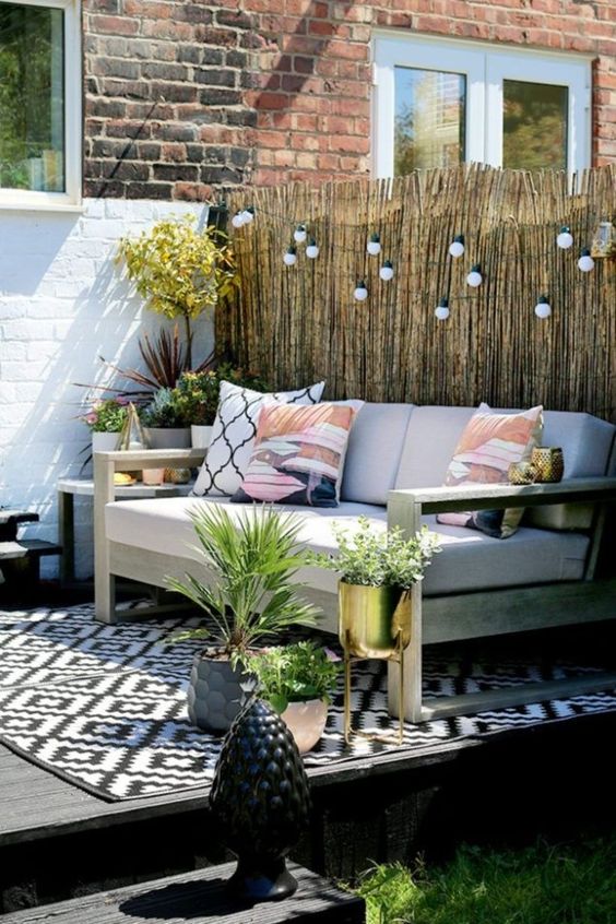 a small deck with a large comfortable sofa, lights over it and lots of potted greenery and blooms