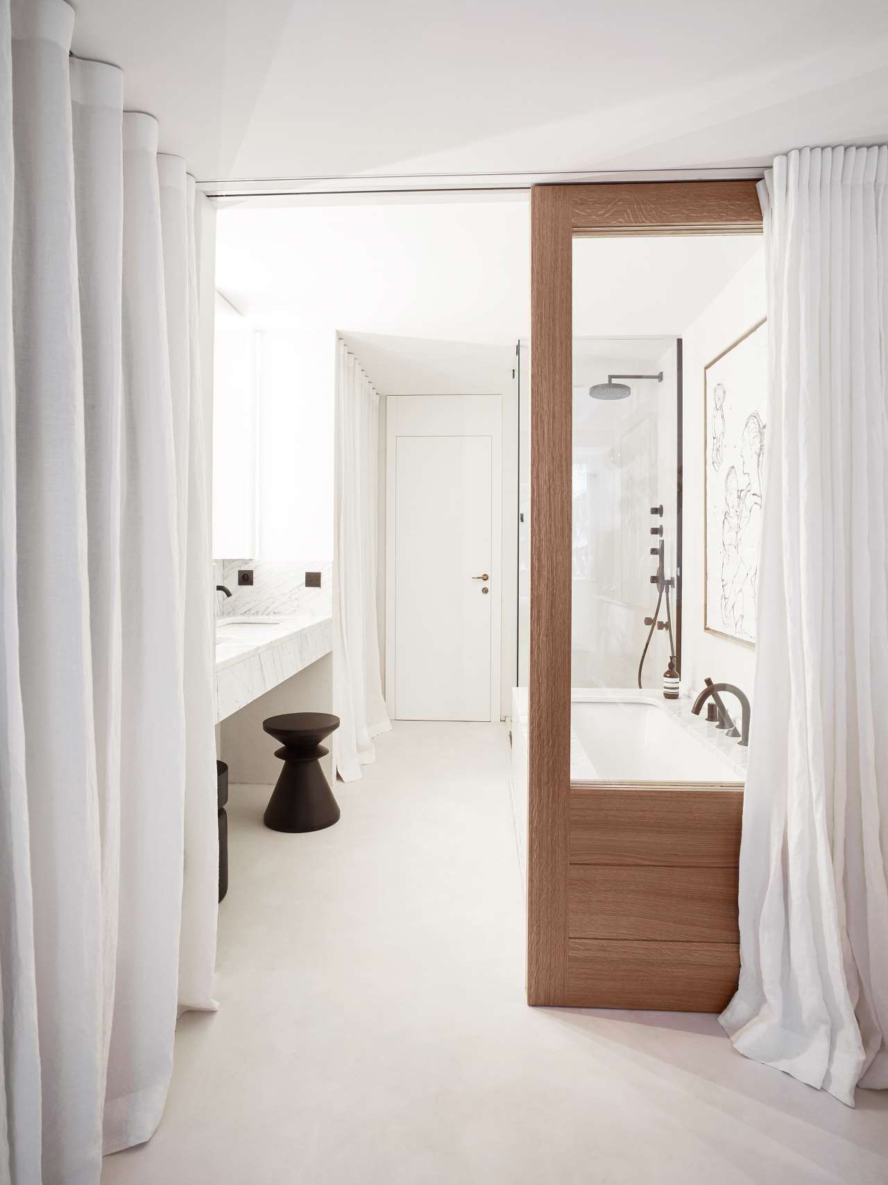 The bbathroom is also white, with a tub, a marble floating vanity, a wooden door and some curtains for privacy