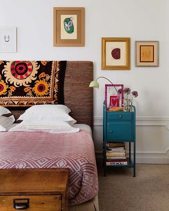 a bright eclectic bedroom with an upholstered bed, a wooden chest, a bight vintage nightstand and abstract art