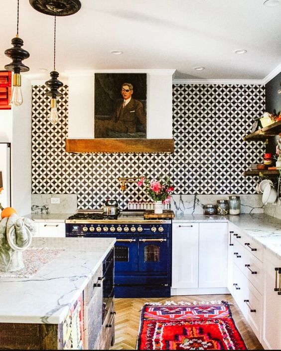 a bright eclectic kitchen with white cabinets, a black and a black and white tile wall, a kitchen island, a bold rug and pendant lamps