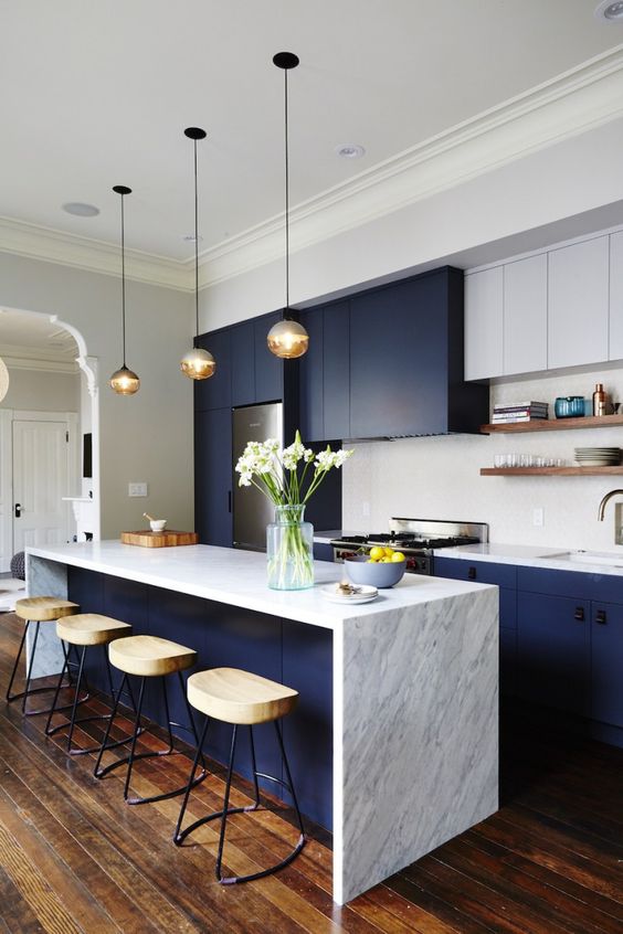 a bright navy and white galley kitchen with a stone countertop, pendant lamps, wooden stools and a white tile backsplash