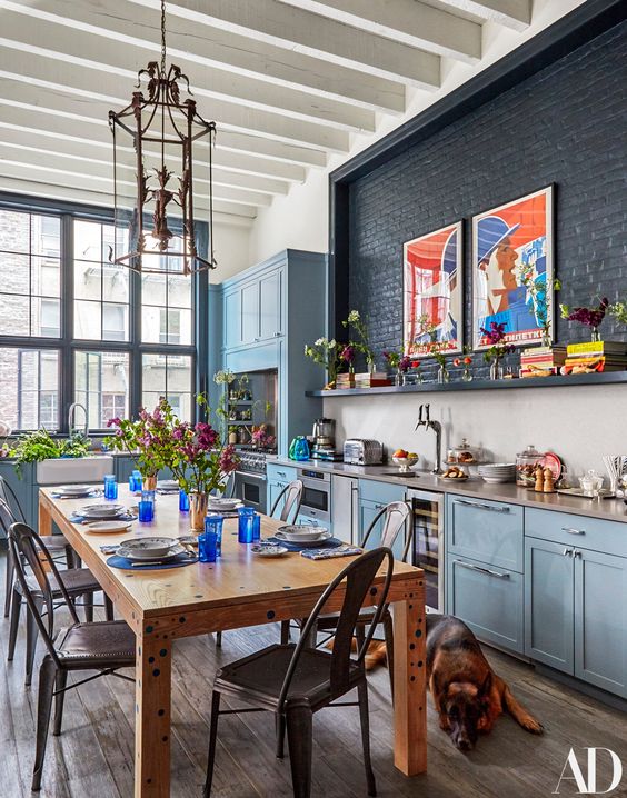 a catchy eclectic kitchen with pastel blue cabinets, a black brick wall, a dining zone with a wooden table and metal chairs, a refined pendant lamp