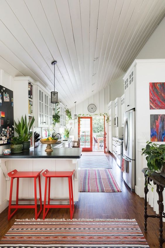 a catchy eclectic kitchen with white cabinets, bold red stools and doors, pendant lamps and potted greenery