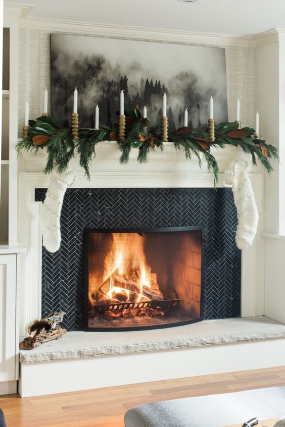 a fireplace surrounded with black herringbone tiles, a white mantel and some greenery and leaves is a stylish and catchy solution