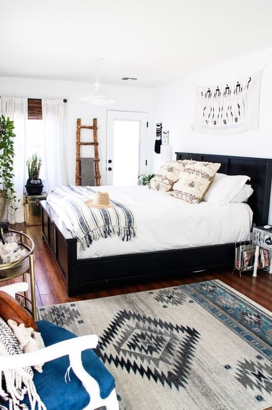 a light filled eclectic bedroom with a dark bed, a rustic ladder for storage, prints, glam tables and potted greenery