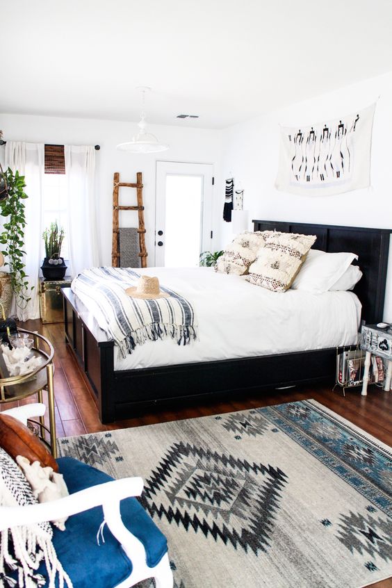 a light filled eclectic bedroom with a dark bed, a rustic ladder for storage, prints, glam tables and potted greenery