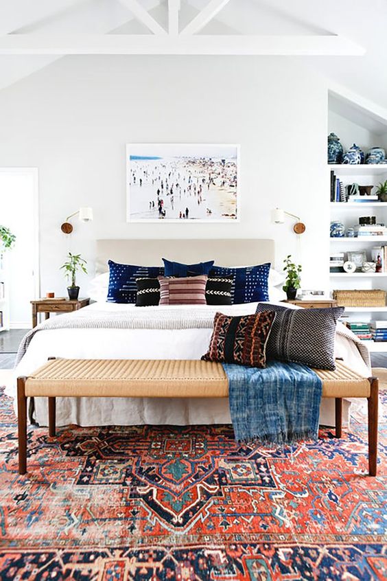 a light filled eclectic bedroom with an upholstered bed, a woven bench, built in shelves, wooden nightstands and potted greenery