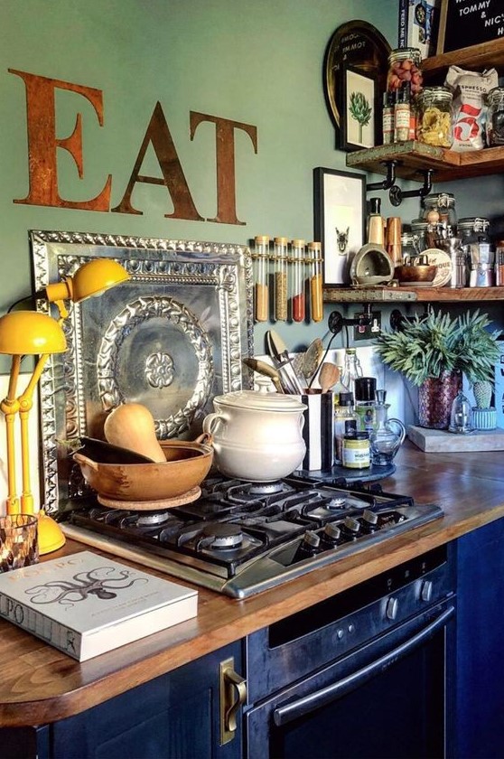 a maximalist kitchen with green walls and navy cabinets, butcherblock countertops, rough wooden shelves and a yellow lamp