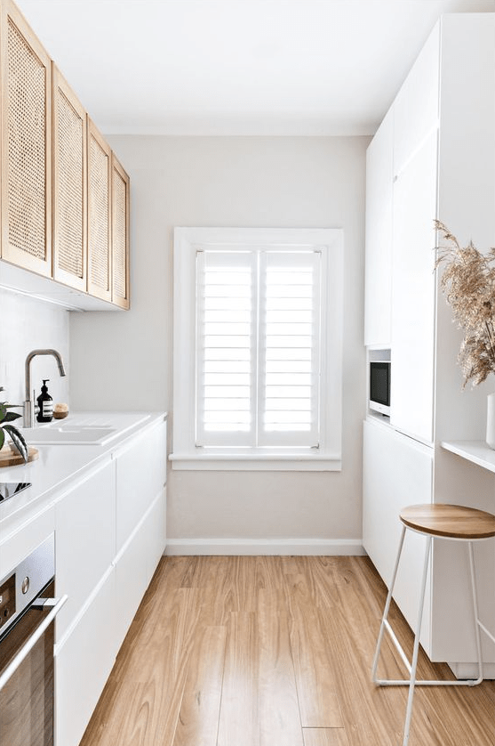 a minimalist white galley kitchen with sleek cabinetry, upper rattan cabinets, a wooden stool and some grass