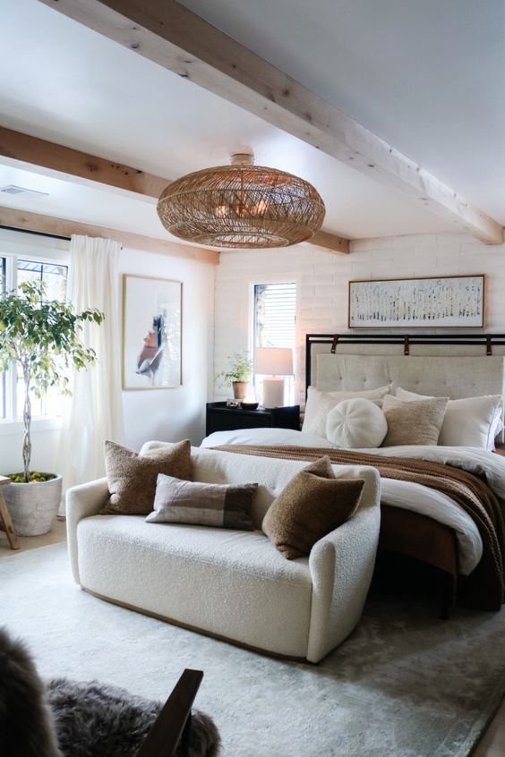 a neutral eclectic bedroom with a white bed, a creamy loveseat with pillows, a woven pendant lamp and some artwork