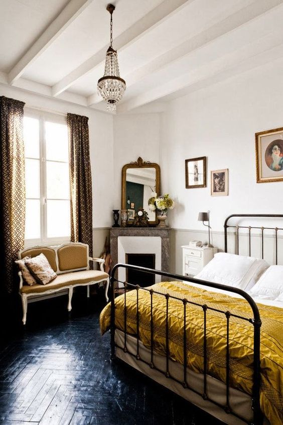 a refined eclectic bedroom with black parquet floor, a metal bed with mustard bedding, a vintage loveseat, a chandelier and a corner fireplace
