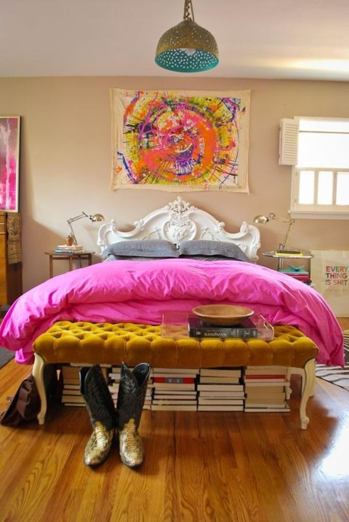 a super bright eclectic bedroom with bold artworks, a mustard tufted ottoman, stacks of books, a refined bed and mismatching nightstands