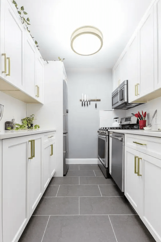 a white galley kitchen with shaker cabinets, white stone countertops, gold handles, a white backsplash and a ceiling lamp