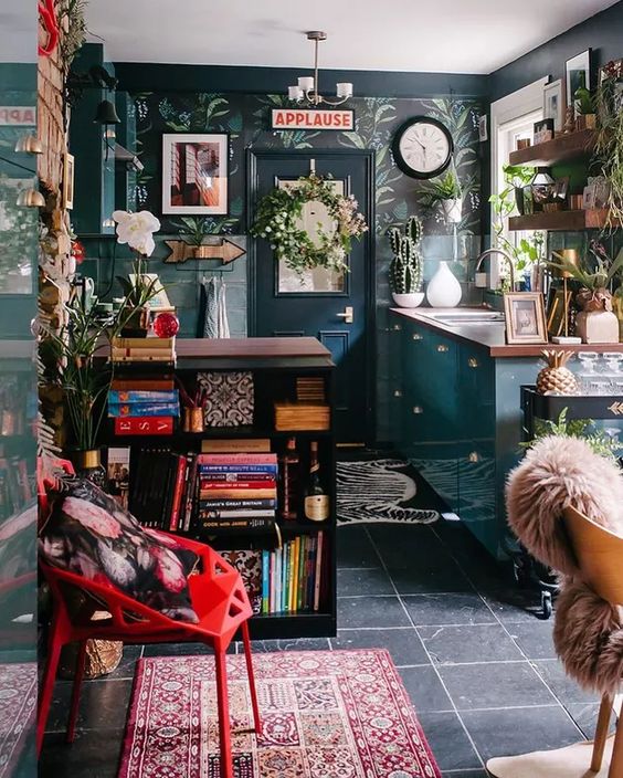 an eclectic and maximalist kitchen with dark wallpaper, teal cabinets, a black bookcase, some shelves and greenery plus a bold rug