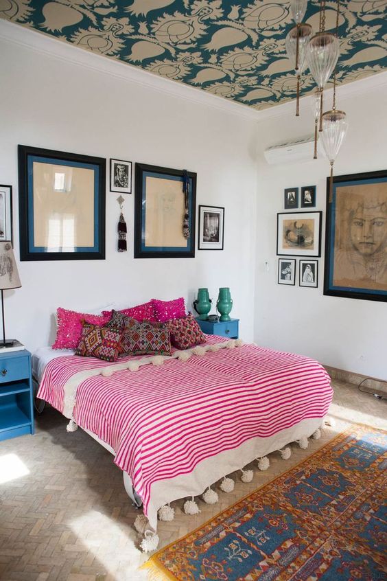 an eclectic bedroom with a wallpaper ceiling, a boho rug and bedding with pompoms, a variety of artworks and retro nightstands