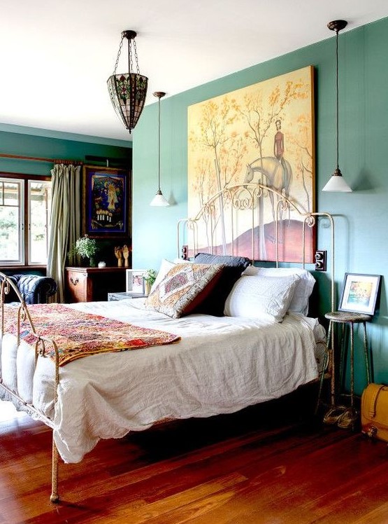 an eclectic bedroom with green walls, a forged bed with colorful and printed bedding, forged stools, a dark-stained desk, a chic chandelier and an oversized artwork
