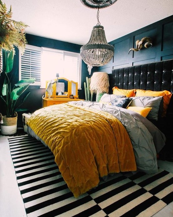 an eclectic bedroom with navy paneled walls, a black upholstered bed, colorful yellow bedding, a yellow vanity, potted statement plants and a beaded chandelier