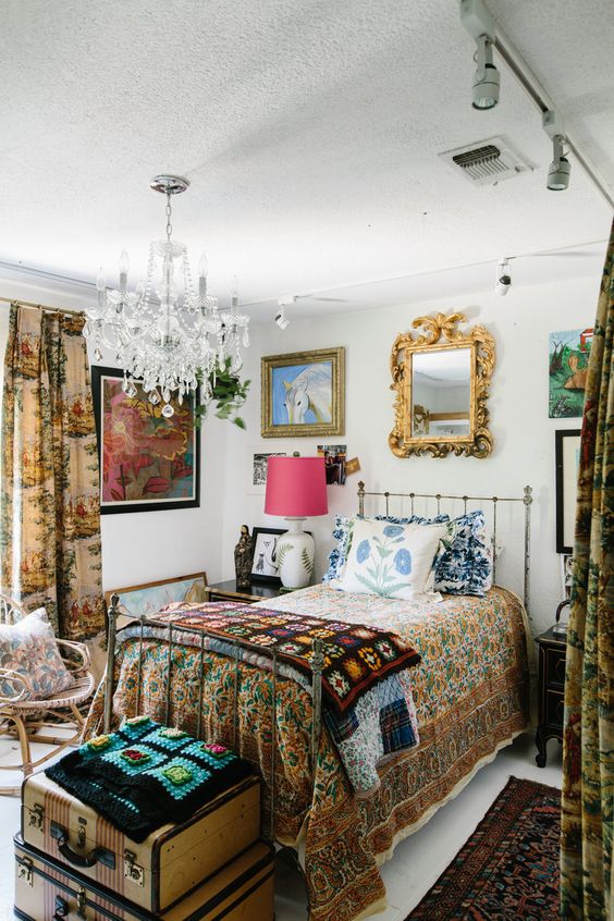 an eclectic guest bedroom with a metal bed and bright textiles, a pouf, a rattan chair, a gallery wall and a crystal chandelier
