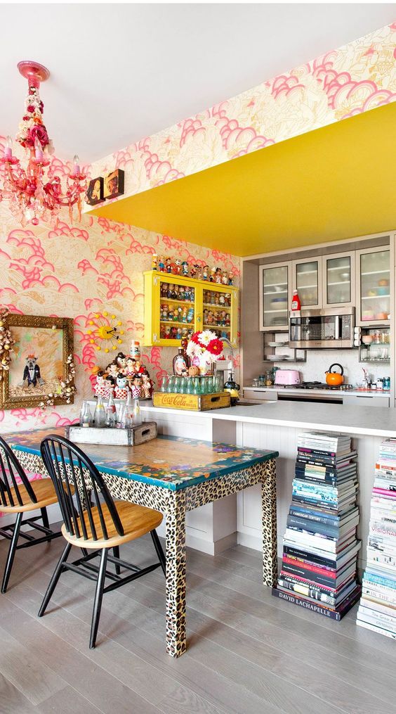 an eclectic kitchen with bold pink wallpaper, a yellow cabinet and a ceiling, a white kitchen island, book stacks, a pink chandelier, a leopard table and stained chairs