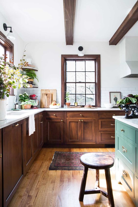 an eclectic kitchen with dark-stained cabinets, a green kitchen island, dark wooden beams and a stool, potted greenery