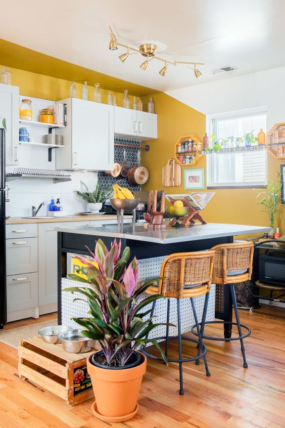 an eclectic kitchen with mustard walls, white cabinets and a black kitchen island, rattan stools, a crate and potted plants