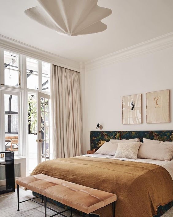 an elegant eclectic bedroom with a bed and a dark headboard, an upholstered bench, some artwork and a pendant lamp