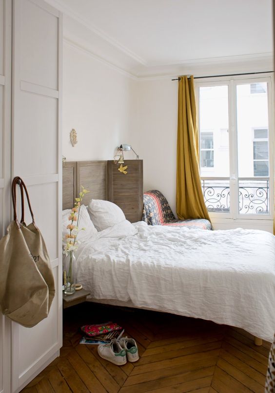 a Paris-inspired chic bedroom with a shutter screen as a headboard, a herringbone floor, mustard curtains and a comfy chair