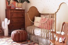 a beautiful boho nursery with a polka dot wall, a bright boho rug, wooden and rattan furniture, a leather ottoman, greenery and a cactus