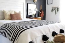 a black and white boho bedroom with a black statement wall, a white bed, a wooden bench, a gallery wall with hats and a tassel blanket