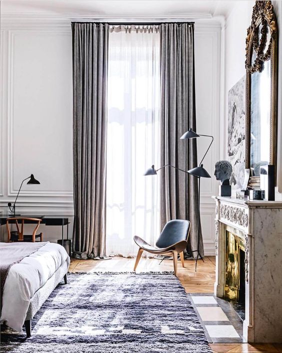 a chic sleeping space with grey textiles, a leather chair, a small desk in the corner, a non-working fireplace and a comfy bed