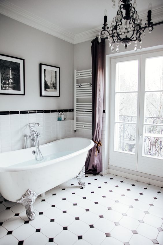 a chic vintage inspired bathroom with a clawfoot bathtub, purple curtains, a crystal chandelier, artworks and black and white tiles