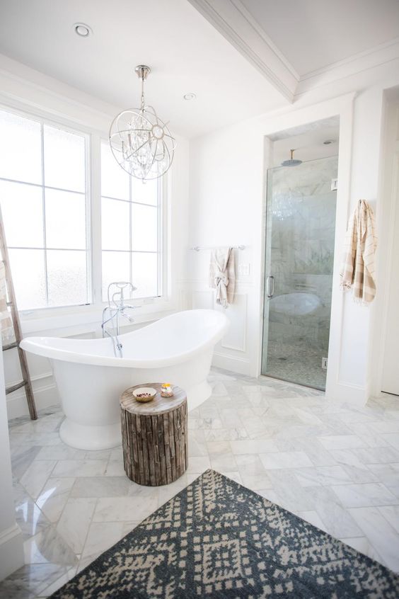 a gorgeous transitional space with marble tiles, a wooden stool, a printed rug, a catchy chandelier and a vintage-inspired tub